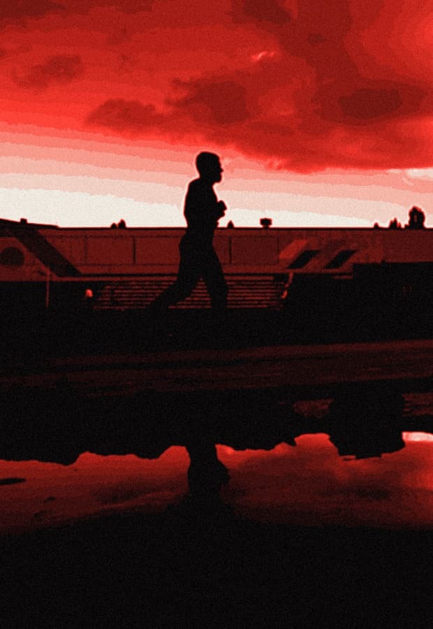 Silhouetted runner alone on an empty stadium track at dusk, red sky mirrored in a rain puddle