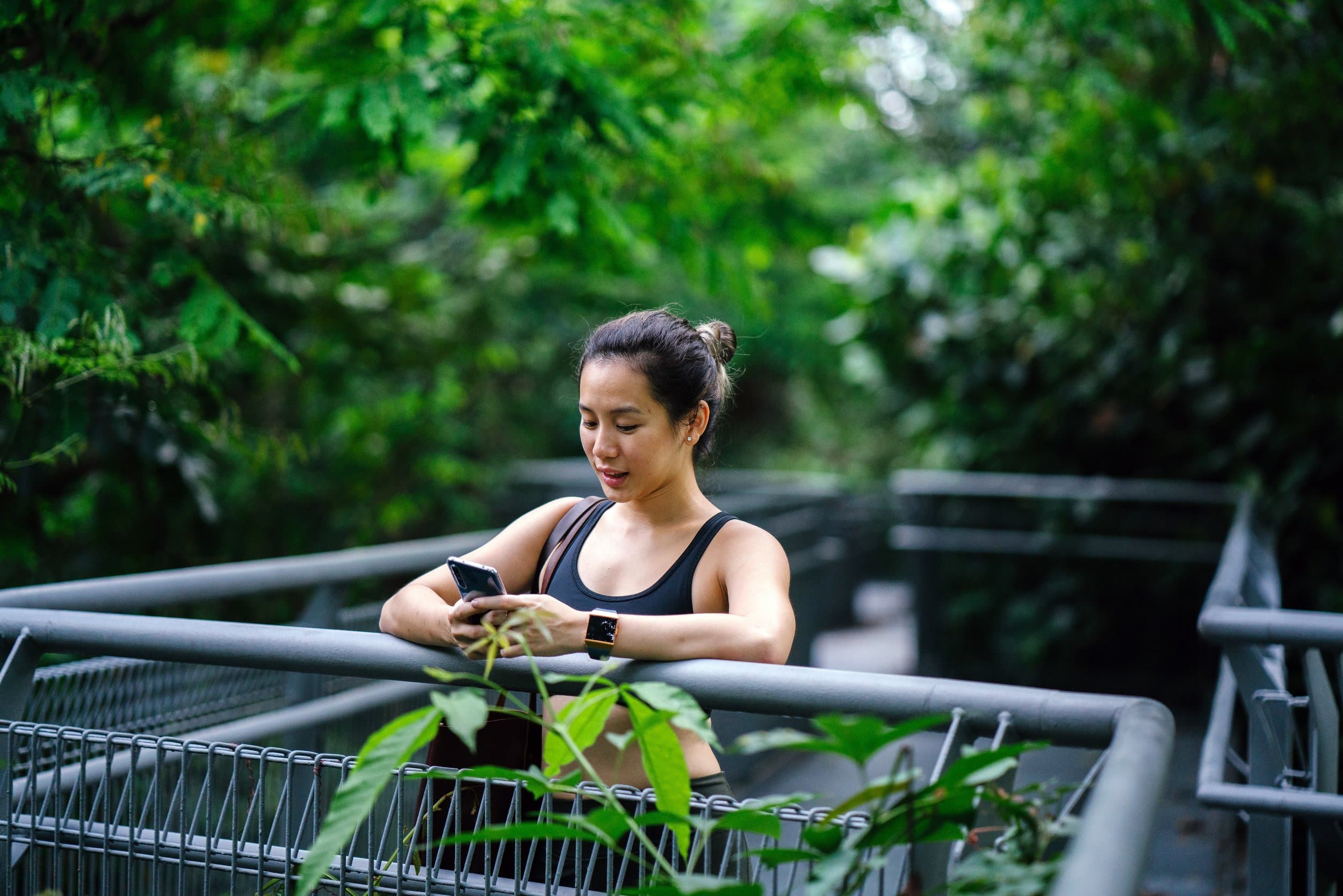 Woman in workout gear leaning on a rail outdoors, checking a fitness app on her phone